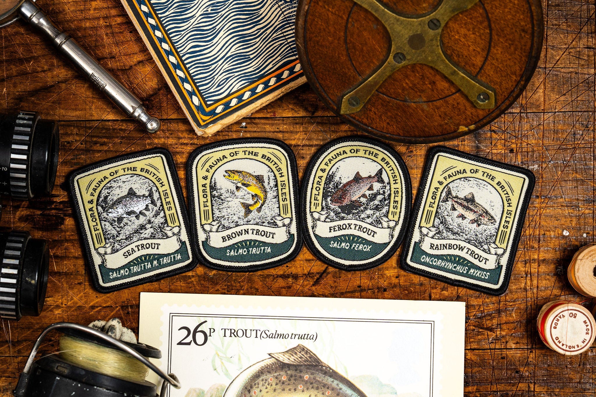 Four vintage-style fishing patch emblems displayed on a wooden tabletop with fishing-themed props, including a reel, magnifying glass, compass, and a trout illustration card.